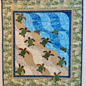 May include: A quilted wall hanging featuring a beach scene with sea turtles. The quilt has a blue border and a central design of blue waves, tan sand, and green and brown turtles. The outer border has a grass pattern.