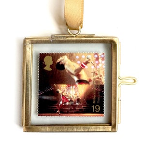 Op de afbeelding: Een gouden metalen frame met een glazen voorzijde toont een postzegel met een foto van Freddie Mercury, de leadzanger van Queen. De zegel is uit de Millennium-serie en heeft de tekst "Millennium Mercury Stamp 19".
