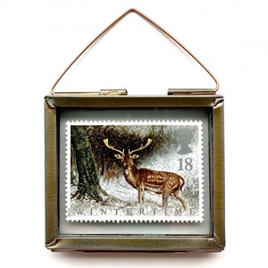 Peut inclure: Un timbre encadré représentant un cerf dans un paysage hivernal. Le timbre est inséré dans un petit cadre rectangulaire en laiton antique avec une attache en fil de cuivre. Le timbre indique "Wintertime" et le chiffre "18".