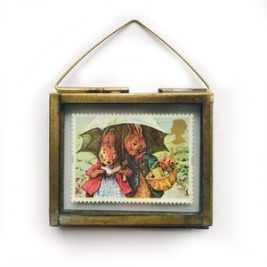 May include: A brass framed picture with a glass front, featuring a vintage postage stamp with two rabbits under an umbrella. The stamp is from Great Britain and features the Queen's profile.