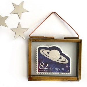 May include: A brass framed picture with a glass front, featuring a vintage Japanese postage stamp with a depiction of Saturn and the text "82 Nippon". The frame has a copper wire hanger.