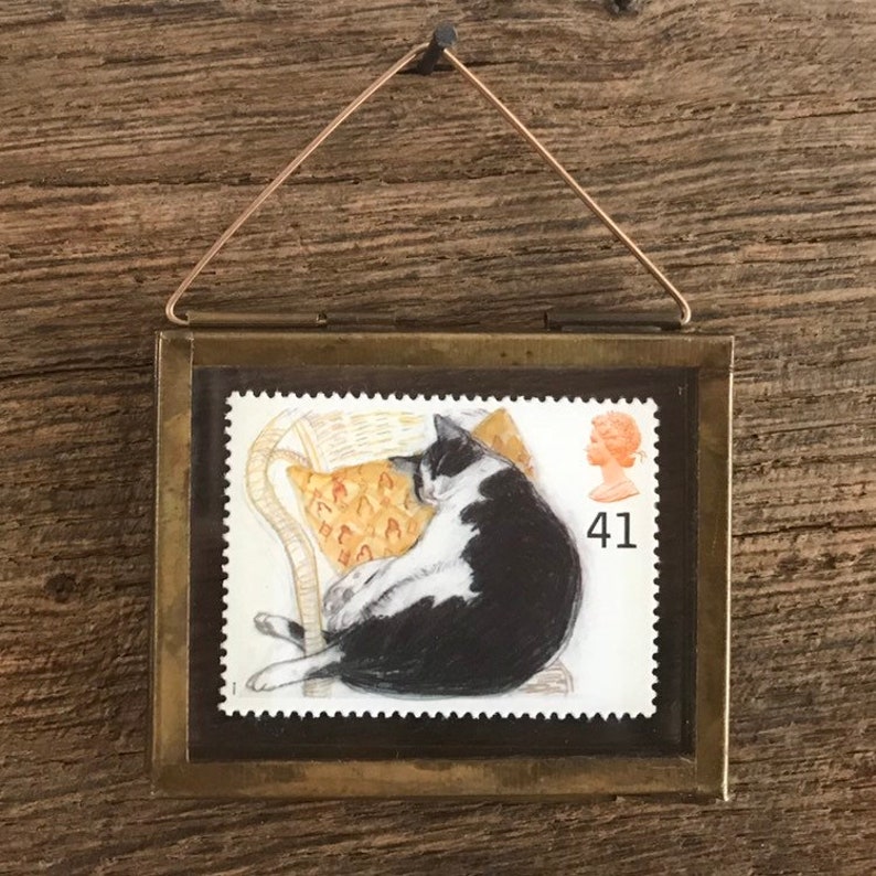 K&ouml;nnte beinhalten: Eine gerahmte Vintage-Briefmarke mit einem schwarz-wei&szlig;en Kater, der in einem Korbstuhl schl&auml;ft. Die Marke stammt aus Gro&szlig;britannien und tr&auml;gt die Nummer 41.