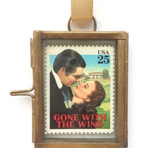 K&ouml;nnte beinhalten: Ein Messingrahmen-Medaillon mit einer Glasfront, das einen Vintage-Briefmarken "Vom Winde verweht" zeigt. Die Briefmarke zeigt eine Szene aus dem Film mit Clark Gable und Vivien Leigh. Die Briefmarke stammt aus den Vereinigten Staaten und hat einen Wert von 25 Cent.
