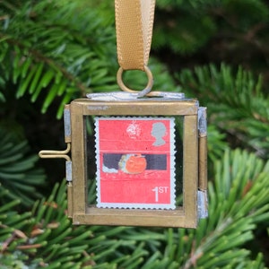May include: A small, square, brass-framed ornament with a vintage postage stamp featuring a red background and a bird illustration. The ornament is suspended by a gold ribbon, perfect for holiday decor.