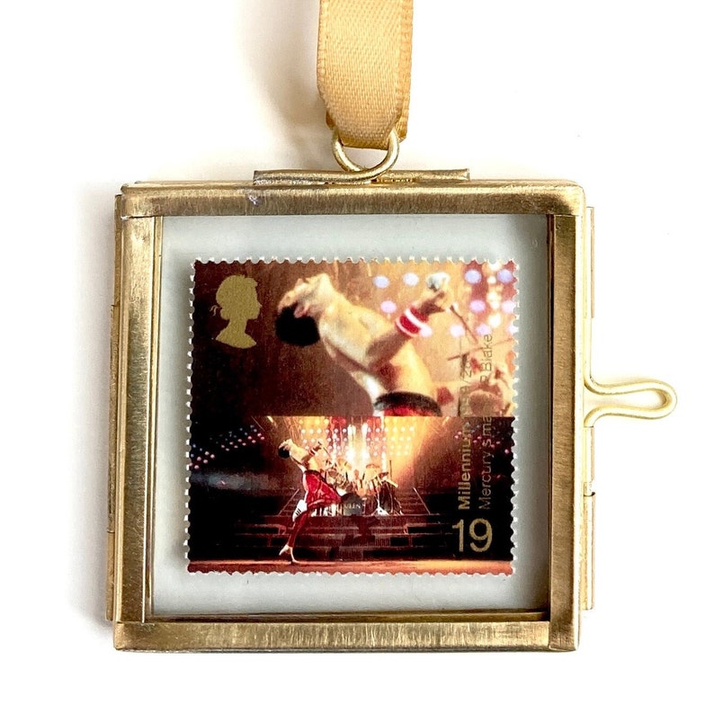 Op de afbeelding: Een gouden metalen frame met een glazen voorzijde toont een postzegel met een foto van Freddie Mercury, de leadzanger van Queen. De zegel is uit de Millennium-serie en heeft de tekst "Millennium Mercury Stamp 19".