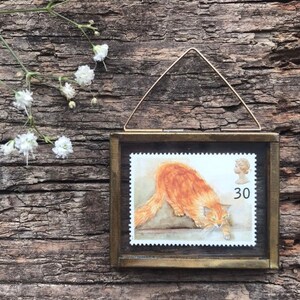 K&ouml;nnte beinhalten: Eine gerahmte Vintage-Briefmarke mit einem roten Kater und einem Nennwert von 30 Pence. Die Marke befindet sich in einem Messingrahmen mit einem Aufh&auml;ngedraht.
