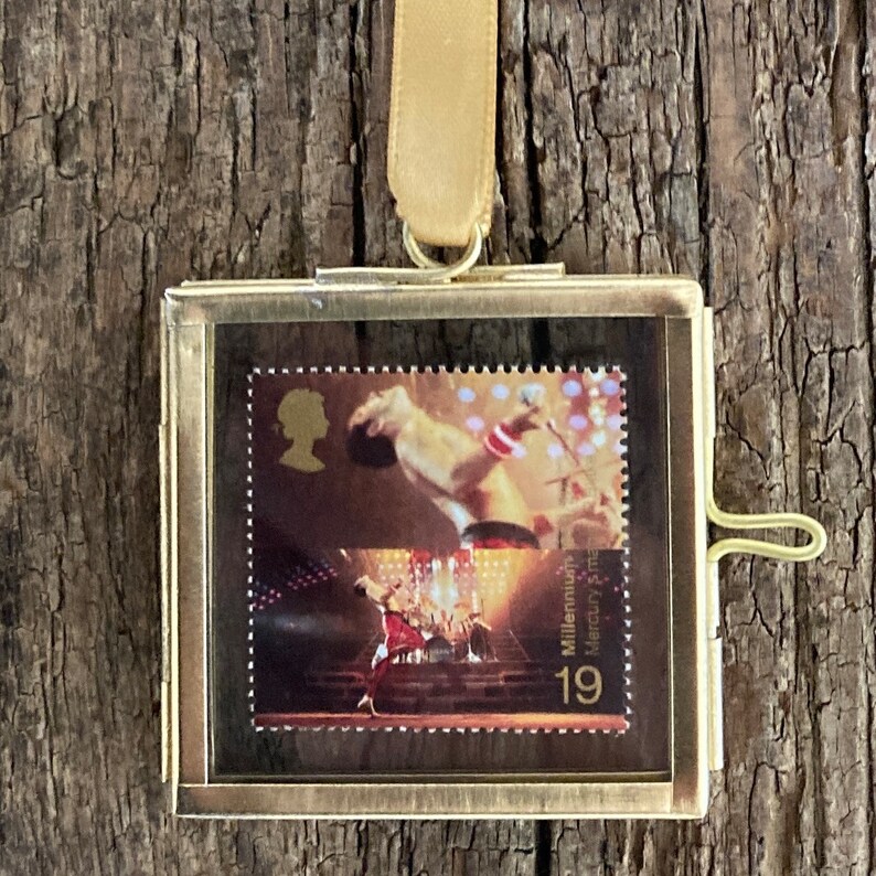 Op de afbeelding: Een goudkleurig ingelijst plaatje van een postzegel met een foto van Freddie Mercury die op het podium optreedt. De postzegel is uit de Millennium Mercury-uitgave en heeft het nummer 19 erop gedrukt.