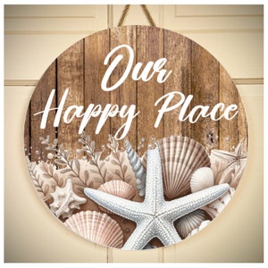 May include: Round wooden sign with the text "Our Happy Place" in white lettering. The sign is decorated with seashells, starfish, and seaweed in shades of brown, beige, and white.