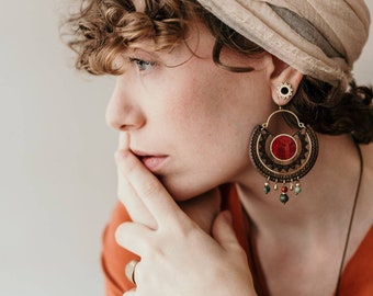 May include: A woman with curly hair wears a beige turban and a pair of large, round, wooden earrings with red and green accents. She is wearing a rust-colored top and gold bracelets.