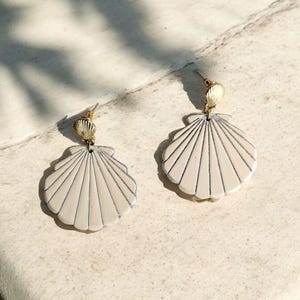 Pode incluir: Um par de brincos em forma de concha. Os brincos são de cor bege claro com linhas cinza escuro e um pino dourado. Os brincos estão sobre uma superfície de cor clara.