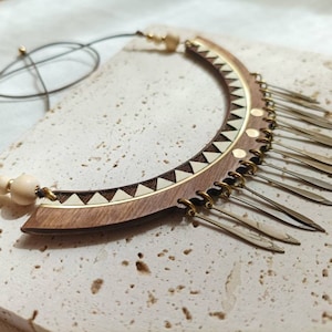 May include: A wooden and metal necklace with a geometric design and a fringe of metal accents. The necklace is brown and gold with white accents.