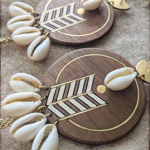 Peut inclure: Une paire de boucles d'oreilles en bois avec un motif géométrique et une frange de coquillages de cauris blancs.