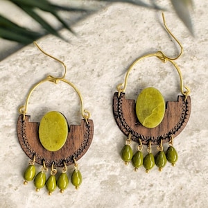 Pode incluir: Um par de brincos artesanais com um aro dourado e uma base de madeira semicircular com detalhes intrincados. Cada brinco tem uma pedra verde oval central e contas verdes penduradas. Brincos para orelhas furadas.