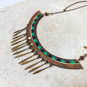 May include: A wooden necklace with a green and black geometric pattern and gold-colored metal accents. The necklace has a fringe of gold-colored metal tassels.
