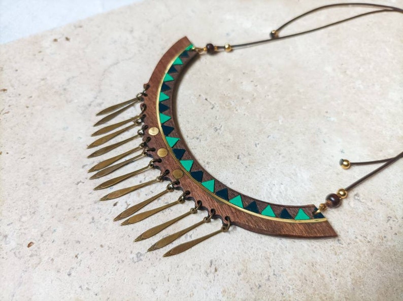 May include: A wooden necklace with a green and black geometric pattern and gold-colored metal accents. The necklace has a fringe of gold-colored metal tassels.