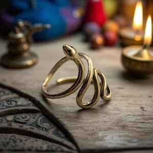 Peut inclure: Une bague serpent dorée avec un design enroulé. La bague présente une tête et un corps de serpent détaillés, reposant sur une surface en bois. L'arrière-plan comprend des bougies allumées et des objets décoratifs.