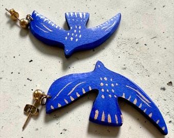 Puede incluir: Un par de pendientes con forma de pájaro azul con detalles pintados en dorado en las alas y el cuerpo. Cada pendiente está unido a un poste de metal dorado con un cierre de mariposa a juego, mostrando un diseño plano y recortado.