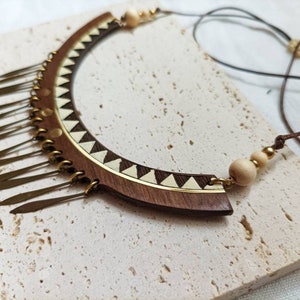 May include: A wooden and brass necklace with a geometric design and a fringe of metal pieces. The necklace is brown and gold and has a white and brown geometric pattern.