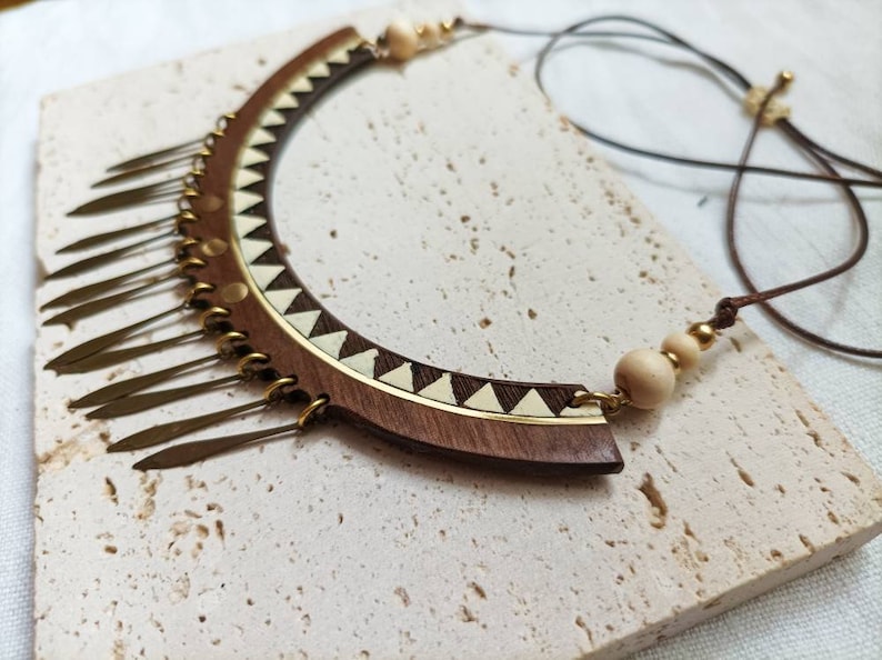 May include: A wooden and brass necklace with a geometric design and a fringe of metal pieces. The necklace is brown and gold and has a white and brown geometric pattern.
