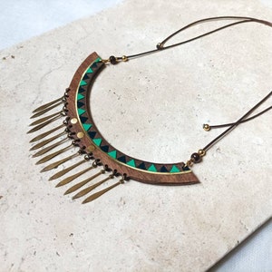 May include: A wooden statement necklace with a geometric design in green, black, and gold. The necklace has a fringe of gold metal accents.