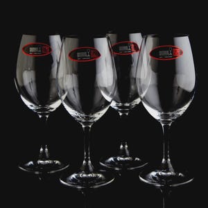 May include: A set of six clear wine glasses with long stems, arranged against a black background. Each glass has a red and white logo sticker. The glasses are designed for wine.