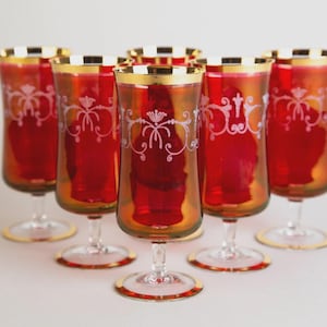 May include: Set of six red-tinted wine glasses with gold rims and bases. Each glass features a white, ornate floral design. The glasses have a tall, slender shape and are arranged on a white surface.