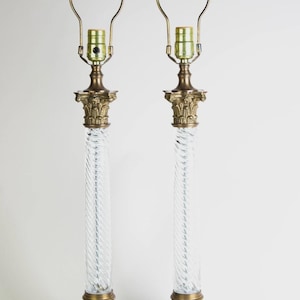 May include: Two table lamps with clear, twisted glass columns and brass-toned bases and accents. The lamps have a classic design with a decorative top and a simple lampshade frame.