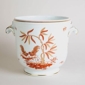 May include: White ceramic planter with a detailed orange and gold design featuring birds and foliage. The planter has two handles and gold trim around the top and base. A decorative piece for plants or home decor.