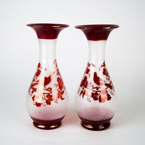 May include: Two frosted glass vases with a red rim and a white background. The vases are decorated with a red and gold floral design and birds.