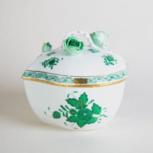 Puede incluir: Un cuenco blanco de porcelana con tapa, diseños florales verdes y ribete dorado. La tapa tiene un asa y adornos de rosas verdes. El cuenco es redondo, probablemente para fines decorativos o de servicio.