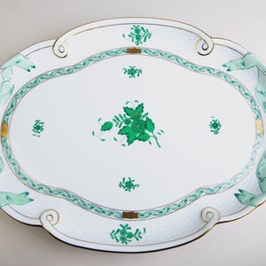 Puede incluir: Una fuente ovalada para servir con base blanca, adornada con diseños florales y cintas verdes. El borde presenta un ribete dorado y asas decorativas. La fuente mide aproximadamente 46 cm de largo y 30 cm de ancho.