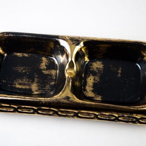 May include: A black and gold ceramic ashtray with two compartments. The ashtray has a decorative border.