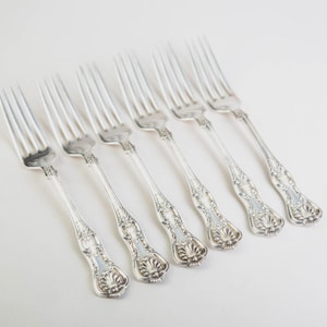 May include: Six silver forks with ornate floral detailing on the handles are arranged diagonally on a white surface. The tines of the forks are visible, and the overall design is elegant and classic.