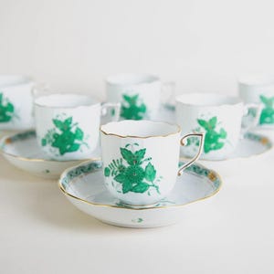 Puede incluir: Un juego de seis tazas y platillos blancos. Cada taza y platillo presenta un diseño floral verde. Los bordes de las tazas y platillos están adornados con oro. Los platillos tienen un borde festoneado con un borde verde y dorado.