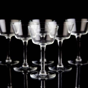 May include: Six clear, stemmed cocktail glasses arranged in a semi-circle against a black background. The glasses have a shallow, wide bowl and a slender stem, reflecting light. The glassware is likely for serving cocktails.