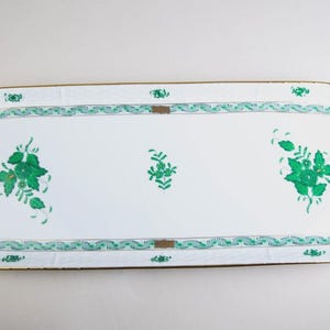 Puede incluir: Una bandeja ovalada blanca con un diseño floral verde y ribete dorado. La bandeja tiene asas en cada extremo y un borde decorativo con un patrón repetitivo. Presenta tres grupos de diseños florales verdes.