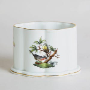 May include: White ceramic container with a scalloped rim and base, adorned with a gold trim. The container features a detailed illustration of two birds perched on a branch with green leaves. The design is centered on the front of the container.