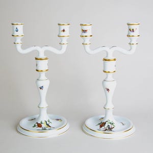 Puede incluir: Dos candelabros de porcelana blanca con detalles dorados y motivos de mariposas y pájaros. Cada uno tiene tres brazos para velas. Las bases presentan pájaros y diseños florales pintados, añadiendo elegancia.
