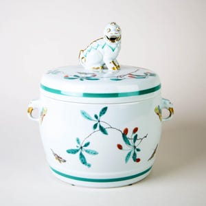 Puede incluir: Un recipiente blanco de cerámica con tapa y diseño decorativo. El recipiente presenta una banda verde alrededor de la base y la tapa, con motivos florales y de hojas. Una pequeña figura blanca y dorada se encuentra sobre la tapa.