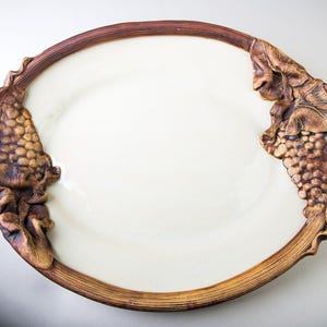 May include: A white ceramic oval serving platter with brown, sculpted grapevine handles. The handles are detailed with leaves and grapes.