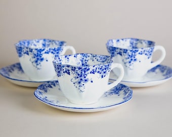 May include: Three white teacups and saucers with a blue floral design. The teacups have a scalloped rim and a curved handle. The saucers have a matching blue floral pattern around the edge.
