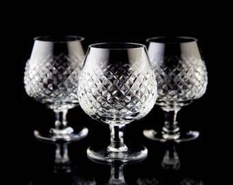 Puede incluir: Tres copas de brandy de cristal transparente con patrones tallados en diamante. Las copas tienen forma de cuenco redondeado, un tallo corto y una base ancha. El fondo negro resalta la claridad y el brillo de la cristalería.