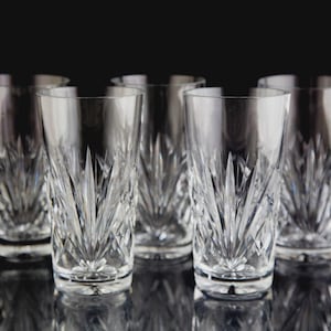 May include: Six clear, cut-glass highball glasses with a starburst design. The glasses are arranged in a semi-circle on a reflective black surface, showcasing their intricate patterns and elegant form.