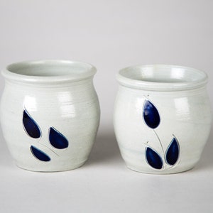 May include: Two white ceramic pots with blue leaf designs. The pot on the left has three blue leaves, while the pot on the right has a single blue leaf with a stem and two smaller leaves.