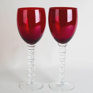 May include: Two ruby red wine glasses with clear, textured stems and bases. The bowls of the glasses are a deep, rich red, reflecting light. The stems have a unique, stacked design. The glasses are set against a plain white background.