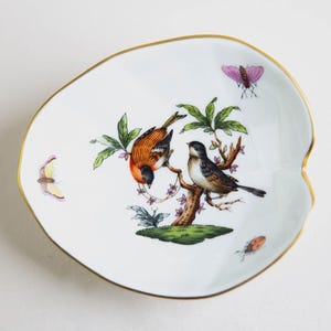 Puede incluir: Plato decorativo blanco en forma de hoja con borde dorado. El plato presenta una colorida ilustración de dos pájaros posados en una rama de árbol, con mariposas y una mariquita. Una pieza encantadora para exhibir o servir.