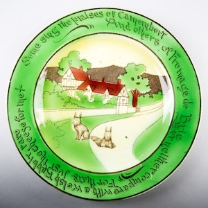 May include: A vintage ceramic plate with a green border and a white center. The plate features a whimsical illustration of a countryside scene with two rabbits and a house. The text around the border reads "Some sing the praises of Camembert And others of Thomage de Brie But neither compare with a welsh Rabbit rare For that's just the cheese for me."