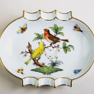 Puede incluir: Plato decorativo ovalado blanco con borde dorado y bordes festoneados. El plato presenta una escena pintada de dos pájaros posados en una rama con hojas verdes y mariposas. Los pájaros son amarillos y marrones.