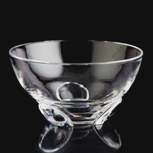 May include: A clear glass bowl with a smooth, rounded interior and a thick, sturdy base featuring three distinct, curved, and molded feet that provide elevation and stability to the overall structure of the transparent vessel.