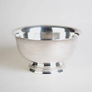 Puede incluir: Un cuenco plateado con una forma ancha y redondeada y una base decorativa. El cuenco tiene una superficie reflectante y se asienta sobre una base de pedestal con un diseño adornado. El cuenco probablemente se utiliza para servir o exhibir.